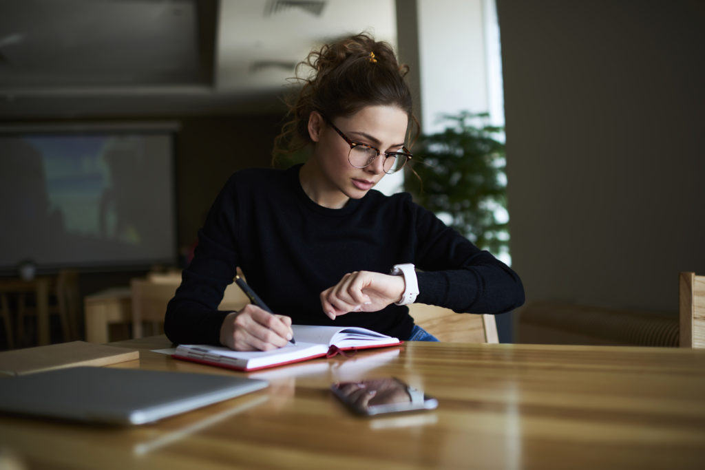 young woman in glasses checking watch while writing in a book