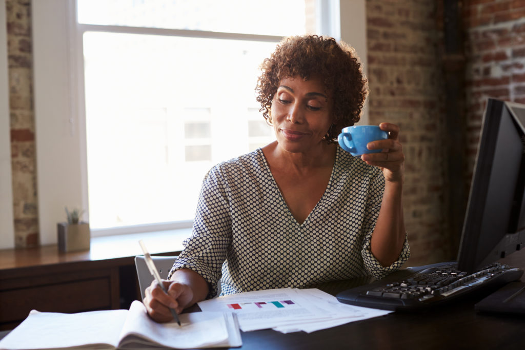mature woman at desk sipping coffee and confidently taking notes on a journal from her computer