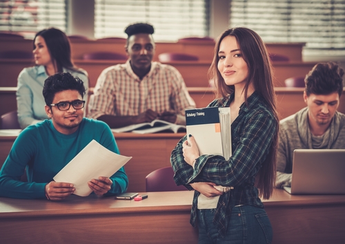 confident woman holding books while standing in front of college classroom